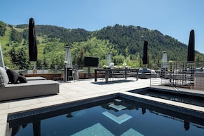 A rooftop pool with a view of a forested mountain, outdoor seating, and a flat-screen TV.