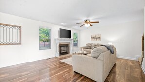 Ein geräumiges Wohnzimmer mit Holzdielen, einem beigen Sofa, einem Kamin und einem Deckenventilator.