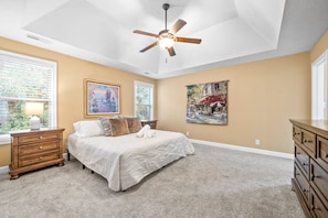 Une chambre à coucher comprenant un lit, un ventilateur de plafond, une table de chevet, une commode et deux fenêtres.