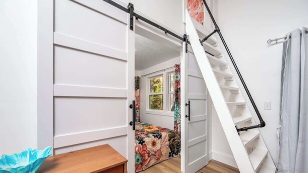 A bedroom with a bed, a wooden nightstand, a white wardrobe, a staircase, and a window with curtains. - Full Bloom Tree House - Stonewall Road (Pittsboro)