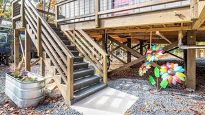 A wooden staircase leading to a deck with a metal railing, a flower sculpture, and a metal planter with plants. - Full Bloom Tree House - Stonewall Road (Pittsboro)