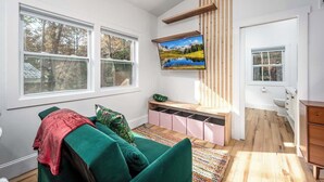 A room with a green sofa, a TV mounted on a wooden wall, a window, and a small kitchen area. - Full Bloom Tree House - Stonewall Road (Pittsboro)