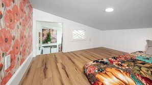 A bedroom with a wooden floor, a colorful bedspread, a white ceiling, and a door leading to another room. - Full Bloom Tree House - Stonewall Road (Pittsboro)