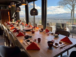 A dining area with a long table set for a meal, large windows with a mountain view, and hanging pendant lights. - Rll (Whitefield)
