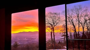 Huone, jossa on suuret ikkunat, joista avautuu näkymä lumiseen maisemaan ja auringonlaskuun.