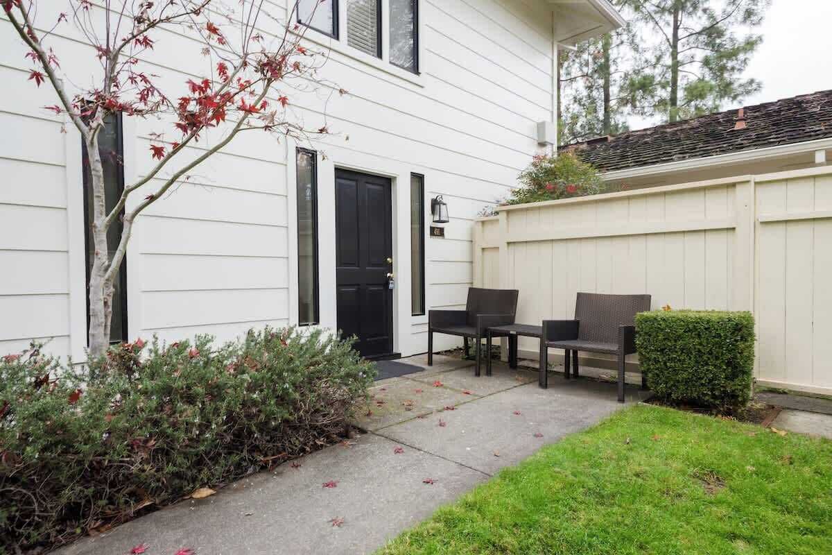 A white house with a black door, a small patio with two chairs, and a hedge.