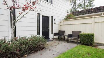 A white house with a black door, a small patio with two chairs, and a hedge.