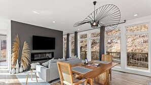 A modern living room with a large TV, grey sofa, wooden dining table, and large windows with a view of trees. - Primrose - Cave Run Lake OR Red River Gorge KY (Wellington)