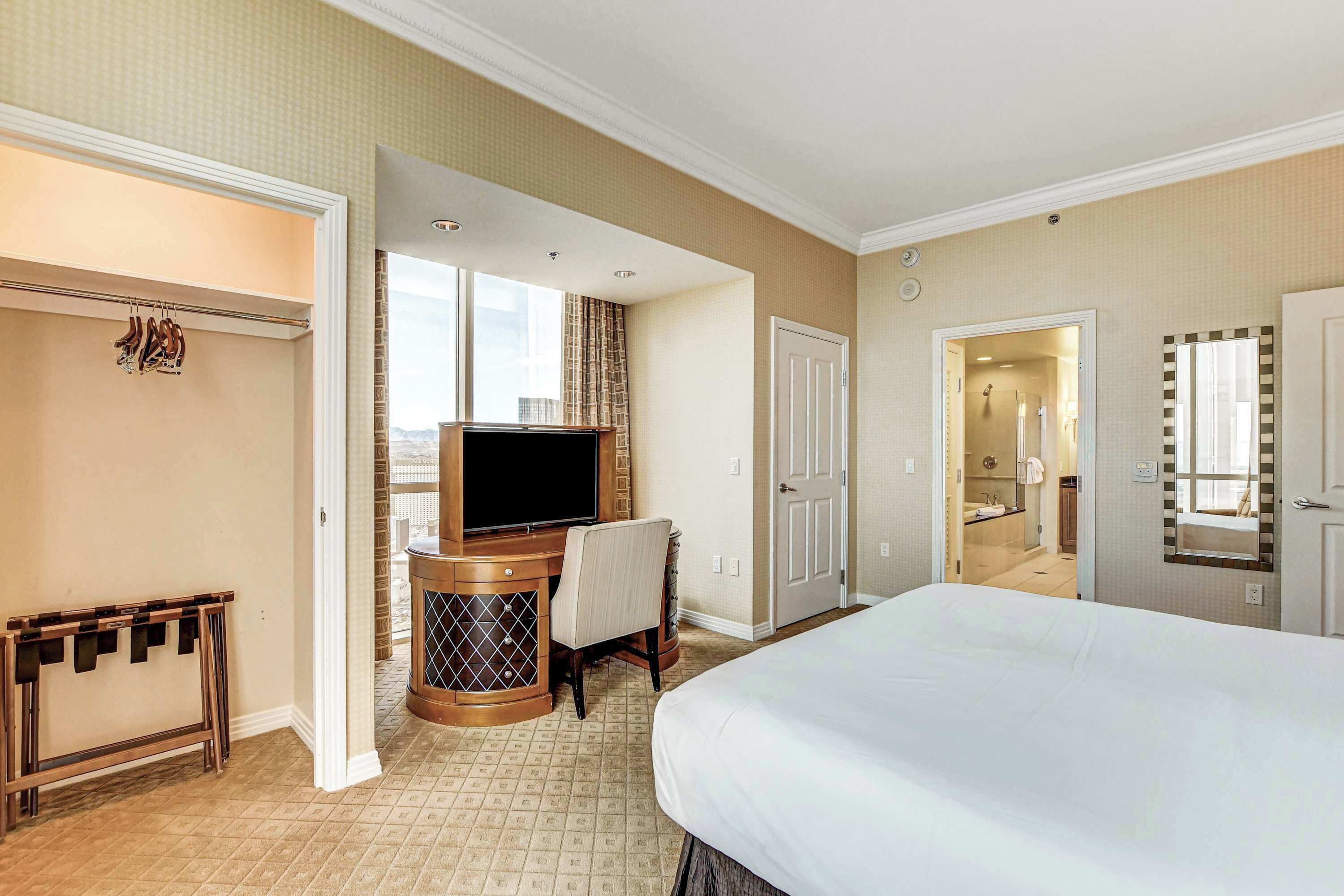 A hotel room with a large bed, a desk with a TV, a chair, and a small table with shoe racks.