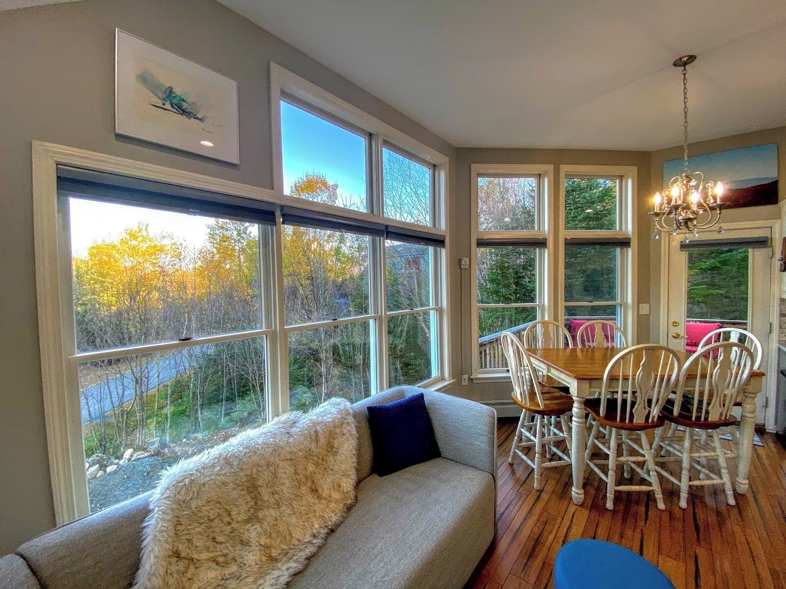 A cozy dining area with a wooden table and white chairs, large windows with a view of trees, and a fluffy white sofa with a blue pillow.