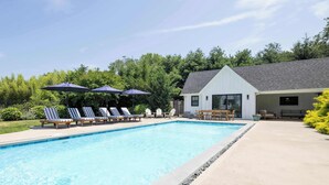 A swimming pool with lounge chairs and umbrellas, a covered seating area, and a building with a sloped roof. - Sandpoint by Rove Travel 6BR Family Home w Pool (Shelter Island)