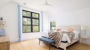 A bedroom with a bed, a bench, a nightstand, a lamp, and a window with curtains. - Sandpoint by Rove Travel 6BR Family Home w Pool (Shelter Island)