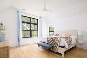 Ein Schlafzimmer mit einem Bett, einer Bank, einem Nachttisch, einer Lampe und einem Fenster mit Vorhängen.