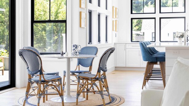A modern living room with a white sofa, a dining area with wicker chairs and a white table, and a kitchen with white cabinets and a blue bar stool. - Sandpoint by Rove Travel 6BR Family Home w Pool (Shelter Island)