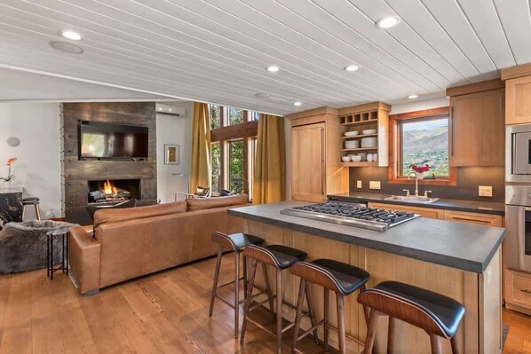 A modern kitchen with a central island, bar stools, wooden cabinets, and a fireplace. - Snowline Lodge 4BR Home With Hot Tub, Pool & BBQ (Aspen)