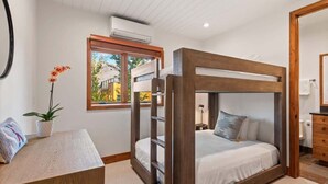 A bunk bed room with a wooden frame, a small desk with a plant, and a window with a view of buildings. - Snowline Lodge 4BR Home With Hot Tub, Pool & BBQ (Aspen)