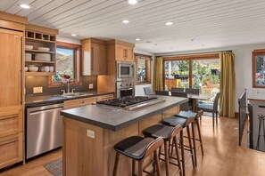 A kitchen with wooden cabinets, a stainless steel dishwasher, a central island with stools, and a view of the outdoors through large windows. - Snowline Lodge 4BR Home With Hot Tub, Pool & BBQ (Aspen)