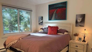 Ein Schlafzimmer mit großem Fenster, einem Bett mit brauner Bettdecke, einem Nachttisch mit Lampe und Bildern an den Wänden.