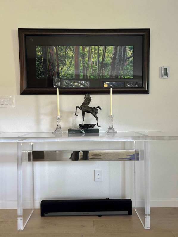A clear glass console table with a mounted flat-screen TV above it, a black Sonos speaker below, and two candles on the table.