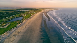 Ein Luftbild einer Küstenregion mit einem langen Sandstrand, einem Golfplatz und einem Gewässer.