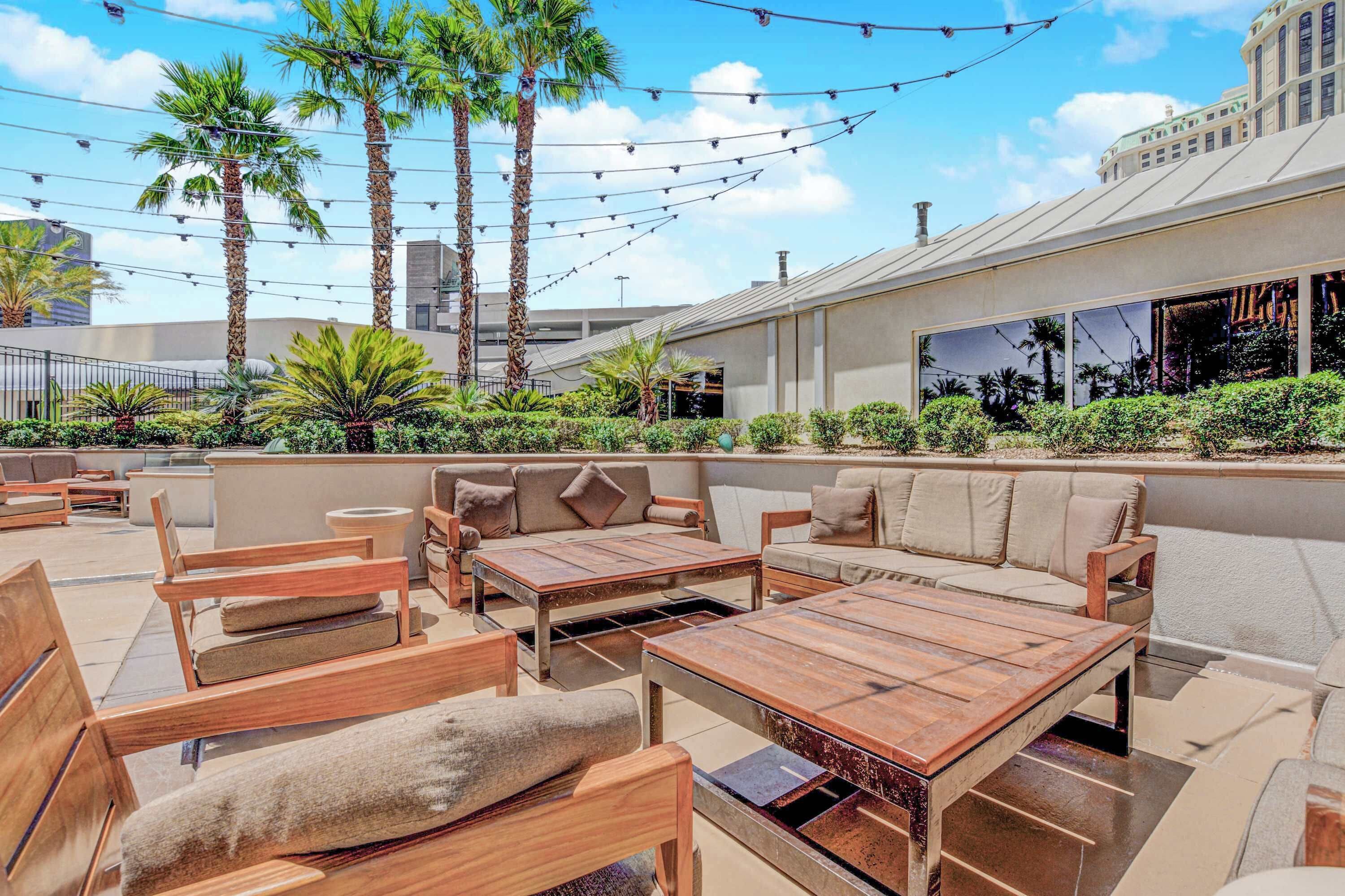An outdoor patio with wooden furniture, palm trees, and string lights.