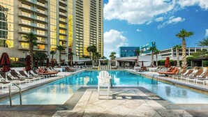 Uma área de piscina de hotel com espreguiçadeiras e guarda-sóis, um edifício alto ao fundo e um céu azul límpido.