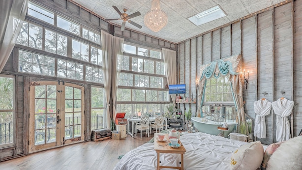 A spacious room with large windows, a bed with white bedding, a wooden ceiling, and a desk with a chair. - The Charleston Treehouse (Wadmalaw Island)