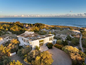 A modern house with a flat roof, surrounded by dense vegetation and a beach view.