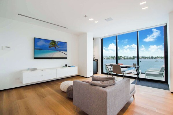 A modern living room with a large TV, a sofa, and a dining area with a view of the water.