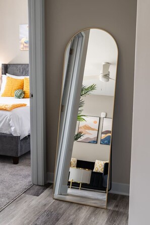 Un dormitorio moderno con un espejo grande, una cama con cabecera gris y una mesita de noche con lámpara.