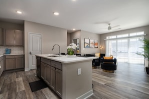 A modern kitchen with a central island, a dining area with a black sofa, and a large window providing natural light. - Lux Haven Whitestown (Whitestown)