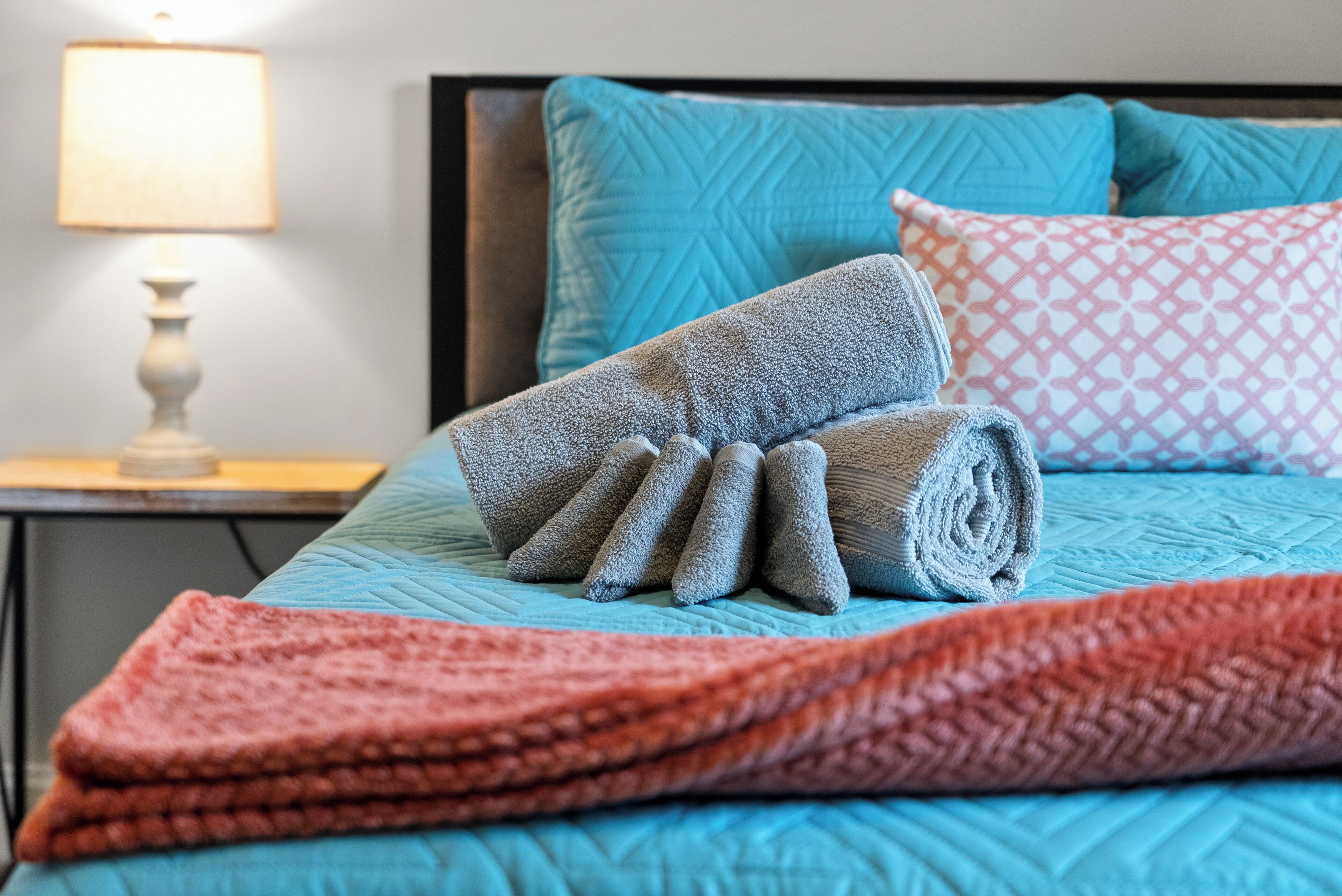 A neatly made bed with a blue quilt, a rolled gray blanket, and a pink patterned pillow.