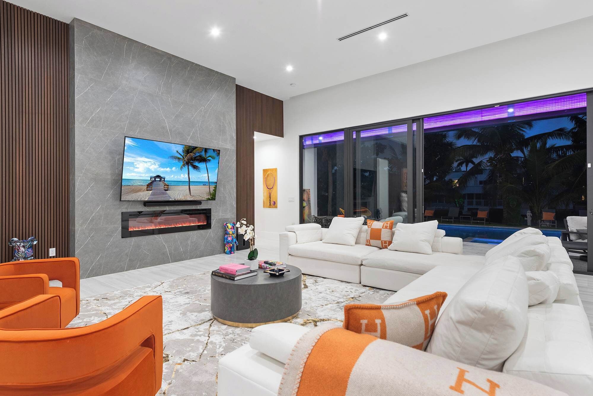 Een moderne woonkamer met een televisie, open haard, witte bank, oranje fauteuil en uitzicht op een zwembad 's nachts.