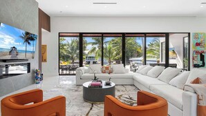 A modern living room with a large sofa, orange chairs, a coffee table, and a view of the beach. - Villa Pesara by Rove Travel Luxury Riverfront (Fort Lauderdale)