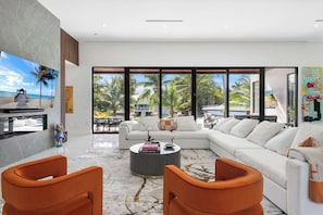 A modern living room with a large sofa, orange chairs, a coffee table, and a view of the beach.
