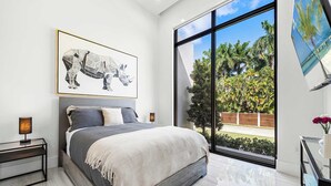 A modern bedroom with a large bed, a nightstand, a framed artwork, and a view of palm trees through large windows. - Villa Pesara by Rove Travel Luxury Riverfront (Fort Lauderdale)