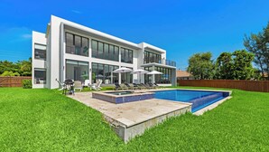 Una moderna casa a due piani con piscina, patio con poltrone a sdraio e ombrelloni, e un prato ben curato.