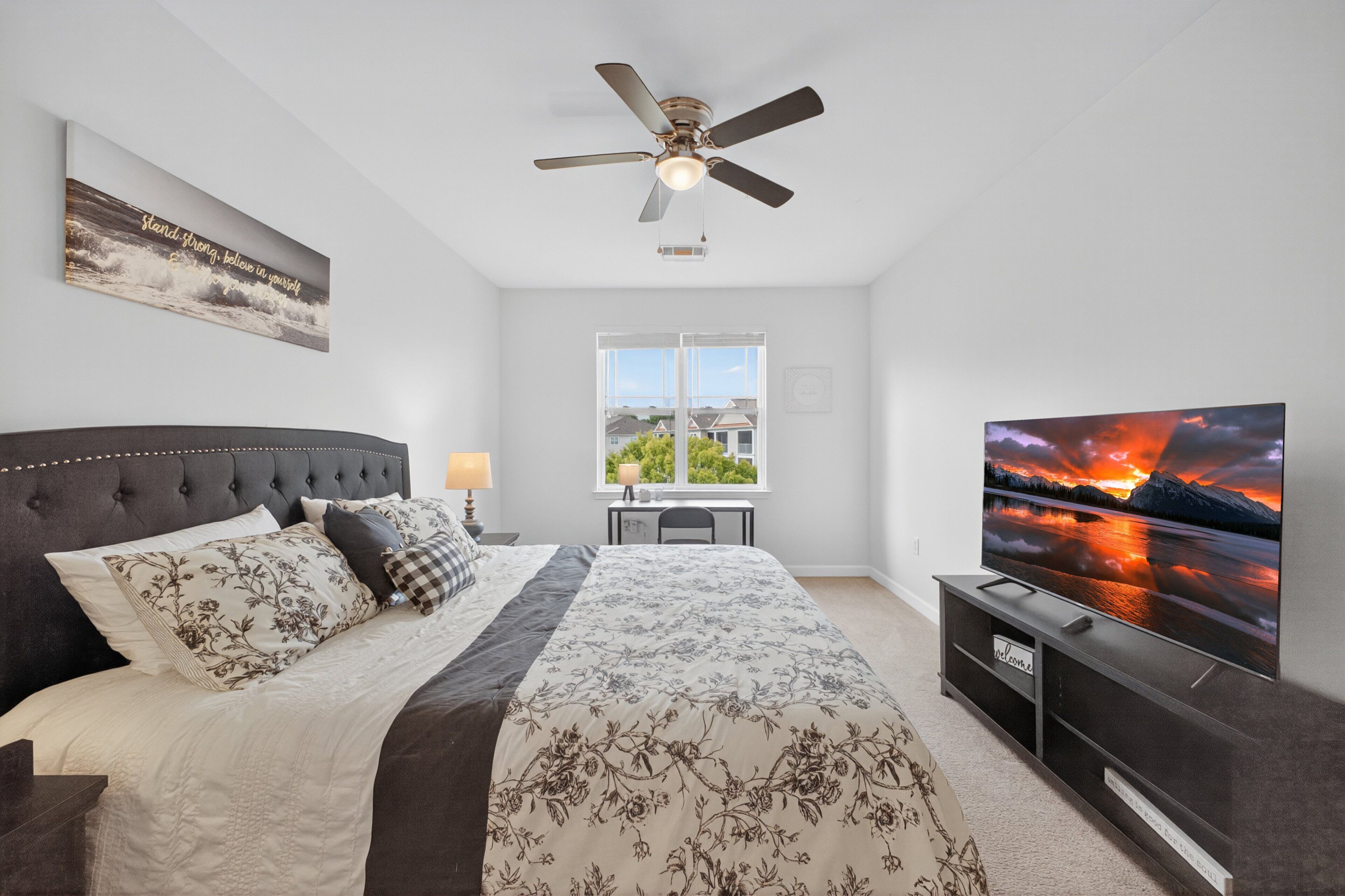 A bedroom with a bed, a ceiling fan, a TV, and a window with a view of houses.