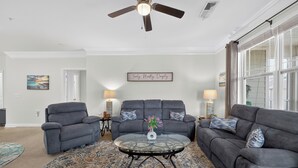 Ein Wohnzimmer mit Deckenventilator, zwei grauen Sofas, einem Glastisch mit einer Vase, einem gemusterten Teppich und zwei Wandleuchten.