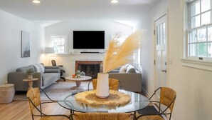 A living room with a fireplace, a flat-screen TV, a sofa, a coffee table, and a glass dining table. - Driftwood Cottage by Rovetravel 3BR Quiet Cottage (East Hampton)