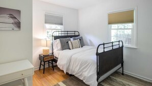 A bedroom with a bed, bedside table, lamp, and two windows. - Driftwood Cottage by Rovetravel 3BR Quiet Cottage (East Hampton)