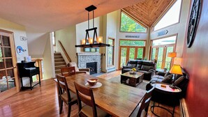 A spacious living area with a fireplace, a dining table with chairs, a sofa, and a large window offering a view of the outdoors.