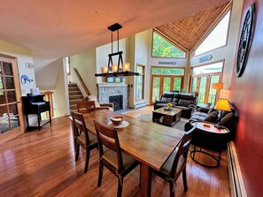 A spacious living area with a fireplace, a dining table with chairs, a sofa, and a large window offering a view of the outdoors.