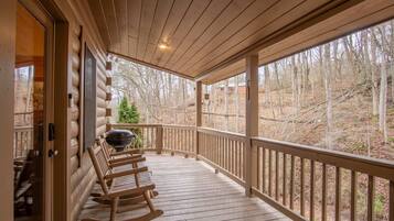 A wooden porch with rocking chairs and a grill, overlooking a forested area.