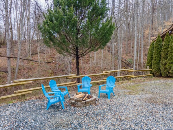 A gravel area with three blue Adirondack chairs, a stone fire pit, and a wooden deck.