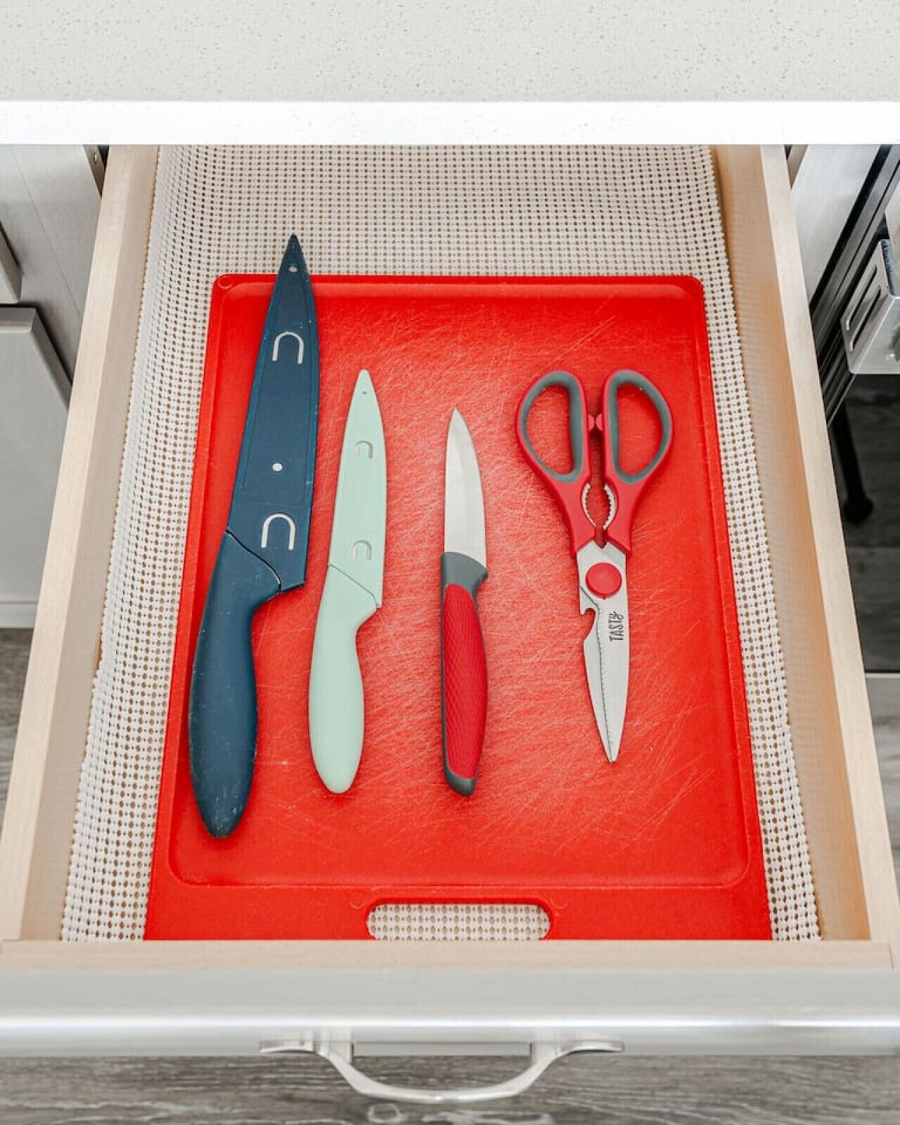 A drawer with a red mat containing three knives, a pair of scissors, and a black knife with a white handle.