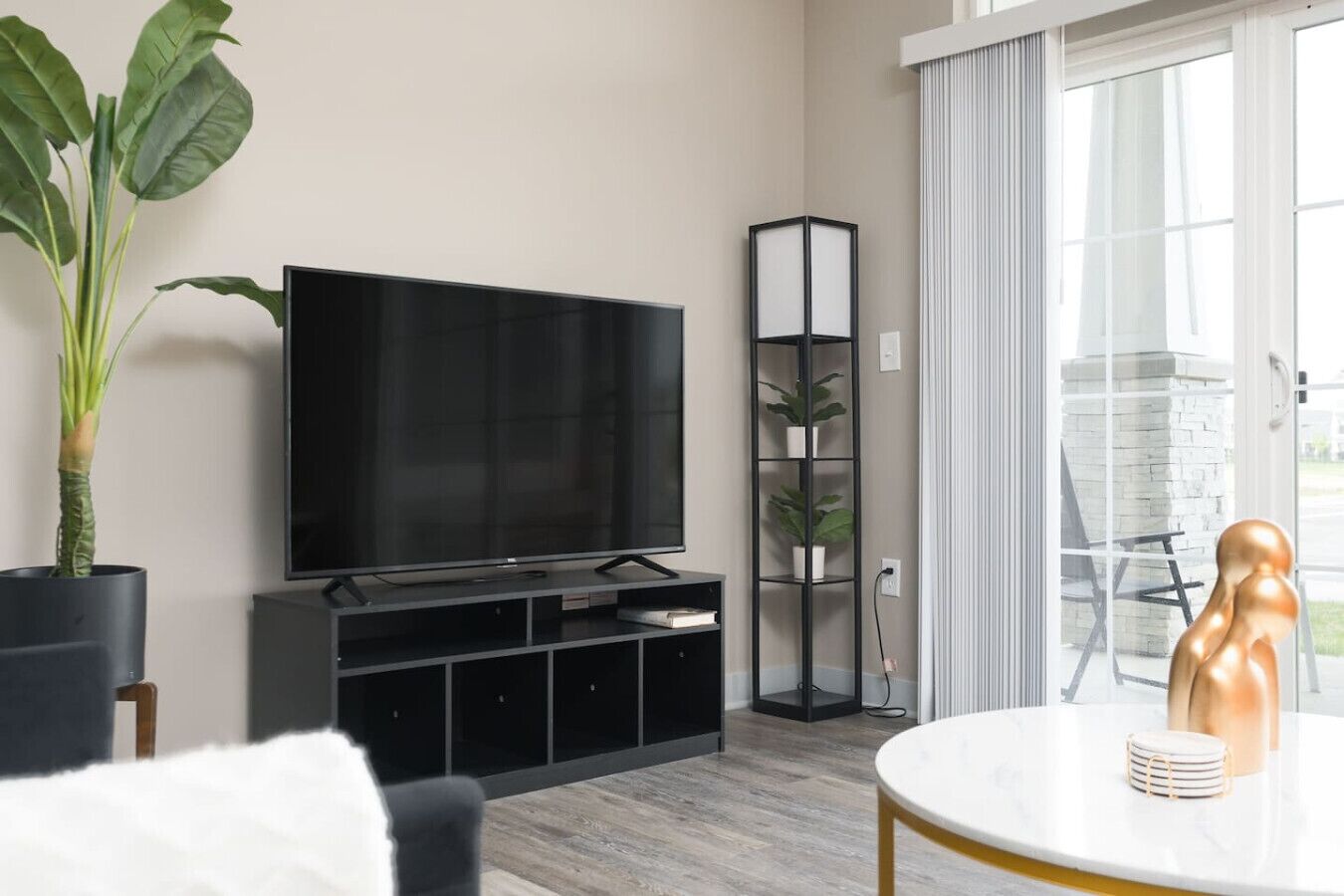 A modern living room with a flat-screen TV, a black entertainment unit, a tall plant stand, and a view of the outdoors through large windows.