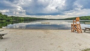 Una playa con un quiosco de salvavidas de madera y un letrero, un banco de madera, y un lago tranquilo con una orilla boscosa bajo un cielo nublado.