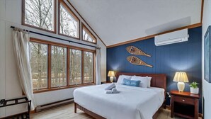 Un dormitorio con un ventanal grande, una cama con ropa blanca, una mesita de noche con lámpara y un aire acondicionado instalado en la pared.