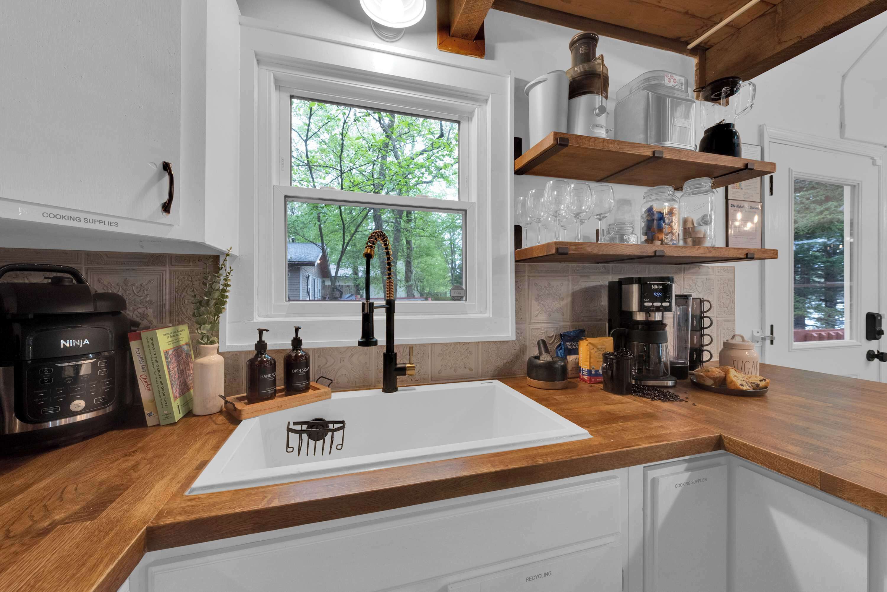 A kitchen with a white sink, wooden countertops, a coffee maker, and a window with a view of trees.